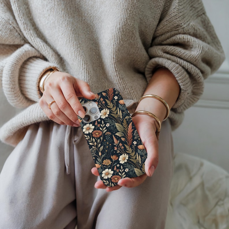 K&ouml;nnte beinhalten: Eine rechteckige Smartphone-H&uuml;lle mit dunklem Hintergrund und einem dichten, komplizierten Muster aus verschiedenen Wildblumen, Gr&auml;sern und Bl&auml;ttern in Creme-, gedeckten Orange-, Braun- und Salbeigr&uuml;nt&ouml;nen, mit Aussparung f&uuml;r die Kamera.