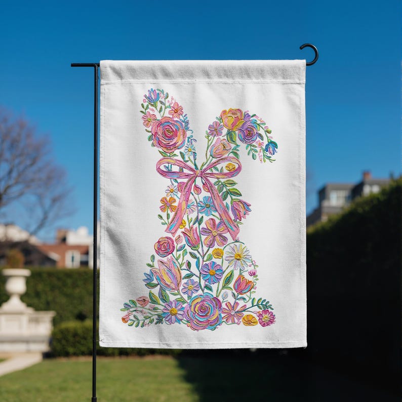 May include: A white garden flag with a floral bunny design. The bunny is made of colourful flowers, including roses and daisies, with a pink ribbon bow. The flag is displayed outdoors on a black metal stand against a blue sky.