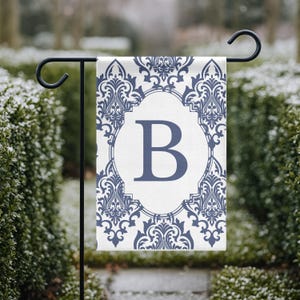Puede incluir: Una bandera de jardín con fondo blanco, adornada con un diseño damasco azul y una gran letra "B" azul. La bandera está colgada de un soporte de metal negro, ideal para la decoración exterior.