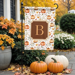 May include: A garden flag featuring a watercolor pattern of pumpkins, leaves, and berries on a white background. A brown square with the letter "B" is centered on the flag. The flag is displayed outdoors with pumpkins and fall foliage.