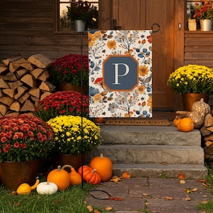 May include: A decorative garden flag with a floral design in shades of orange, blue, and yellow, featuring the letter "P" in a navy blue frame. The scene includes pumpkins, mums, and firewood, creating a fall theme.