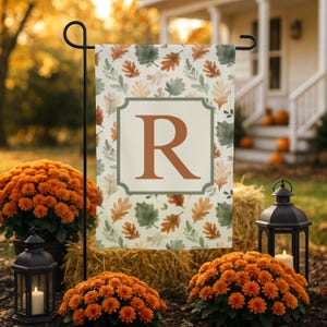 Puede incluir: Una bandera de jardín rectangular con un patrón repetitivo de hojas de otoño en tonos naranja, verde y beige. Un marco cuadrado central con borde verde salvia contiene una letra R mayúscula grande y llamativa en color naranja.