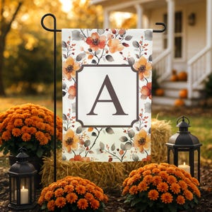 May include: A rectangular garden flag featuring a white background with a pattern of orange and peach flowers and grey leaves. A central dark brown square frame contains a large, bold, dark brown capital letter A in a serif font.