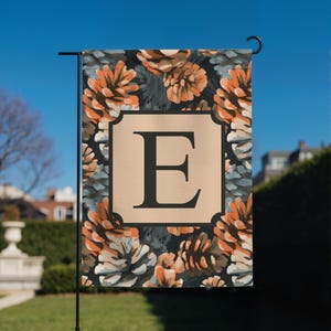 Puede incluir: Una bandera de jardín rectangular con un patrón repetitivo de piñas naranjas, marrones y grises sobre un fondo oscuro. En el centro, un cuadrado beige con borde negro muestra una letra E mayúscula grande y negra en fuente serif.