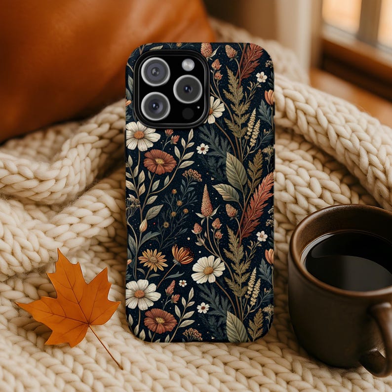 K&ouml;nnte beinhalten: Eine Smartphone-H&uuml;lle mit dunkelblauem Hintergrund und einem wiederkehrenden Muster aus verschiedenen Wildblumen, darunter wei&szlig;e G&auml;nsebl&uuml;mchen, rostfarbene Bl&uuml;ten und gr&uuml;nes Blattwerk. Die H&uuml;lle hat eine Aussparung f&uuml;r eine Dreifachkamera.