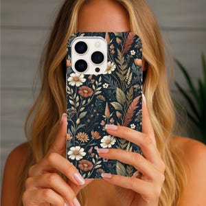 K&ouml;nnte beinhalten: Eine Smartphone-H&uuml;lle mit dunkelblauem Hintergrund und einem wiederkehrenden botanischen Muster aus wei&szlig;en G&auml;nsebl&uuml;mchen, rostfarbenen Bl&uuml;ten sowie verschiedenen gr&uuml;nen und braunen Pflanzenteilen, darunter hohes Gras und bebl&auml;tterte St&auml;ngel.