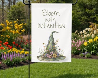 May include: A white garden flag with the text "Bloom with Intention" in green lettering. The flag features a watercolor illustration of a witch's hat adorned with colorful flowers and a bee. The flag is displayed on a black metal stand in a garden setting.