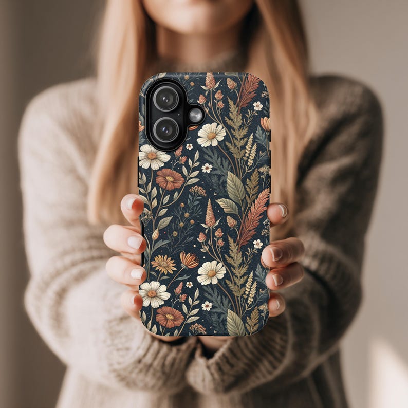 K&ouml;nnte beinhalten: Eine dunkel marineblaue Smartphone-H&uuml;lle mit einem sich wiederholenden botanischen Muster aus wei&szlig;en G&auml;nsebl&uuml;mchen, rostfarbenen Blumen sowie verschiedenen gr&uuml;nen und braunen Bl&auml;ttern. Die H&uuml;lle hat eine Aussparung f&uuml;r eine Doppelkamera.
