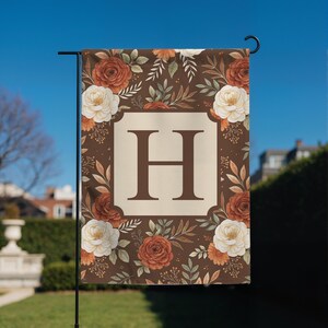 Puede incluir: Una bandera de jardín rectangular con fondo marrón oscuro y un patrón de rosas color óxido y crema, hojas verdes y pequeñas ramitas. Un marco cuadrado de color crema en el centro contiene una letra H mayúscula grande de color marrón.