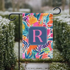 Puede incluir: Una colorida bandera de jardín con un estampado floral en tonos rosa, naranja, azul y amarillo. La bandera presenta un cuadrado azul marino con la letra "R" en rosa. La bandera está colgada en un soporte de metal negro.