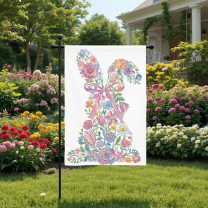 May include: A white garden flag with a floral bunny design. The bunny is made up of colourful flowers, including roses and daisies, with a pink ribbon bow. The flag is displayed on a black metal stand in a garden.