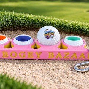 Peut inclure: Un porte-balle de golf rose avec le texte BOGEY BAZAAR gravé en jaune. Il comporte quatre découpes circulaires contenant trois balles de golf blanches à l'intérieur coloré et une balle blanche avec un logo Mulligan Martini et un mousqueton.