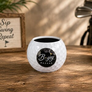 Puede incluir: Una maceta esférica blanca con una textura de hoyuelos que recuerda a una pelota de golf. La parte frontal presenta un logotipo circular negro con el texto blanco "Bogey BAZAAR" y un pequeño icono de bandera de golf. El interior es negro.