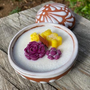 May include: A small, round ceramic bowl with a white, sugary surface, decorated with purple and yellow floral-shaped sweets. A decorative, brown and white patterned ceramic lid sits behind the bowl. The bowl is on a weathered wooden surface.