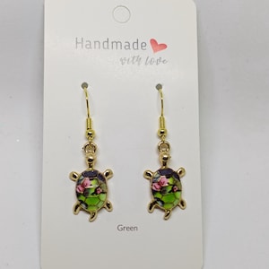 Peut inclure: Boucles d'oreilles dorées en forme de tortue avec des motifs floraux verts, roses et violets. La carte de support indique "Handmade with love". Le mot "Green" est imprimé en dessous. Bijou artisanal.