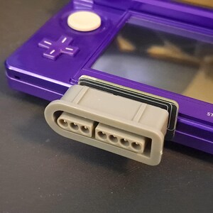 Pode incluir: Close-up de um console de jogos portátil Nintendo 3DS roxo com um adaptador de cartucho cinza conectado. O adaptador possui vários conectores de pinos. O console possui um direcional, botões e uma tela.