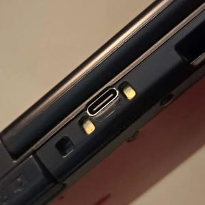 May include: Close-up of a black and red electronic device, featuring a USB-C port. The port is silver, surrounded by gold-colored accents. The device has a metallic gold trim.
