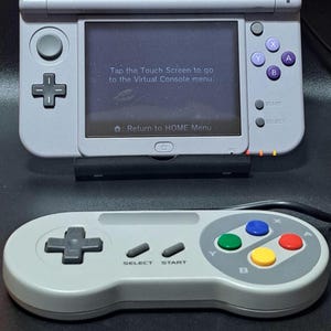May include: A grey Nintendo 3DS handheld console and a matching controller. The 3DS screen shows text: "Tap the Touch Screen to go to the Virtual Console menu." The controller has coloured buttons.