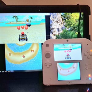 May include: A white Nintendo 2DS handheld gaming console displaying a racing game, alongside a laptop and a tablet showing the same game. The game features a kart racing theme with a beach setting and cartoon characters.