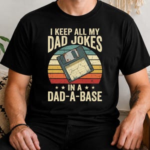 Op de afbeelding: Zwart t-shirt met een retro design met een diskette en de tekst "I KEEP ALL MY DAD JOKES IN A DAD-A-BASE". Het ontwerp bevat een achtergrond in zonsondergangstijl met gele, oranje en rode strepen.