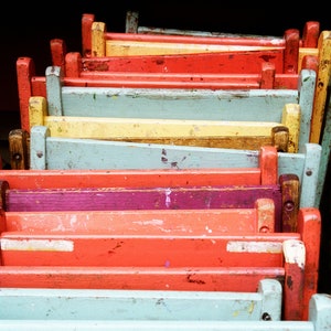 May include: A stack of colorful, weathered wooden chairs. The chairs are painted in a variety of colors, including red, orange, yellow, blue, and purple. The wood grain is visible through the paint, giving them a rustic look.