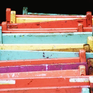 May include: A stack of colorful wooden chairs with various shades of blue, yellow, red, and purple paint. The chairs are stacked in a staggered pattern, creating a visually appealing display.