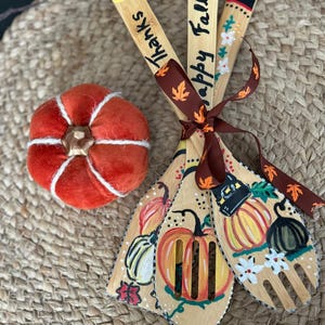 May include: A set of three wooden kitchen utensils with hand-painted fall-themed designs, including pumpkins and the words "Thanks" and "Happy Fall." A small, orange, fabric pumpkin is also present. The utensils are tied with a brown ribbon.