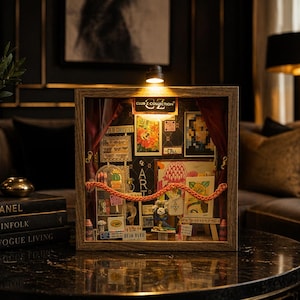 May include: A wooden shadow box display with various art pieces, including drawings and postcards, arranged behind a red curtain. The display is lit by a small overhead light and features the text "ART".