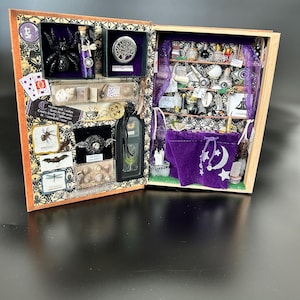 May include: A decorative wooden box, open to reveal a miniature witchy scene. The box is filled with tiny bottles, a black spider, playing cards, and a purple velvet bag with a crescent moon and stars. The box is decorated with patterned paper and small trinkets.