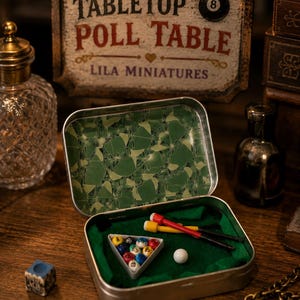 May include: A miniature tabletop pool table set inside a metal tin. The set includes a white triangular rack with small numbered billiard balls, a white cue ball, and two miniature pool cues with red and yellow tips on a green felt surface.