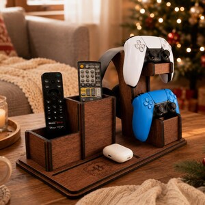 May include: A dark brown wooden organiser stand featuring two vertical compartments for remote controls, a central support for gaming controllers, and a flat base with a small white wireless earbud charging case resting on the surface.