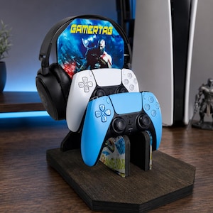 May include: A dark wood gaming stand featuring a vertical display panel with the text GAMERTAG and a character graphic. The stand holds a pair of black over-ear headphones, a white wireless controller, and a light blue wireless controller.
