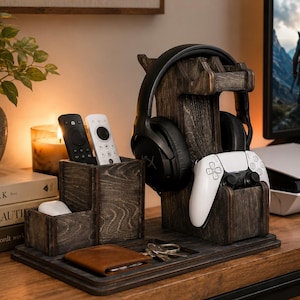May include: A dark stained wooden desk organiser featuring a vertical stand for headphones and a game controller, a dual-compartment holder for remote controls, and a flat tray base for small items like a brown leather wallet and metal keys.