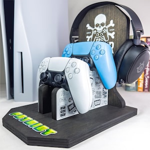 May include: A black wooden gaming controller and headset stand with a white and blue controller and black headset. The stand has a skull and crossbones logo and the word "PATRIOT" in green and yellow.