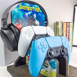 May include: A gaming setup featuring a headset, two game controllers, and a stand with the word "GAMERTAG". The controllers are white and blue. The stand has a colorful design with the words "GAME ON".