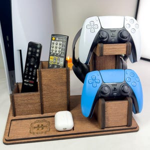 Puede incluir: Un soporte organizador de juegos de madera marrón oscuro con dos compartimentos verticales para mandos, un estante de dos niveles para dos controladores de juego y una base con un estuche de auriculares blanco y un logo B grabado.
