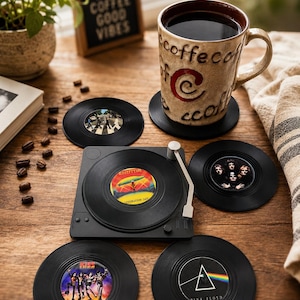 May include: A set of black circular drink coasters designed to resemble vinyl records, featuring iconic album art from bands like Led Zeppelin, Pink Floyd, and Kiss. One coaster is placed under a ceramic mug, while others surround a square record player.