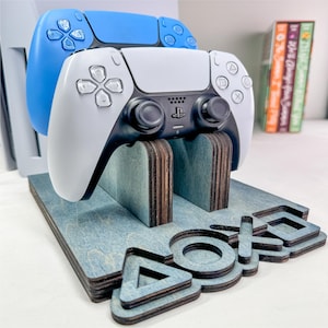 以下が含まれることがあります： 青と白のゲームコントローラーが2つ置かれた、青とグレーの木製ゲームコントローラー用スタンド。スタンドには、PlayStationのロゴが重ねてデザインされています。