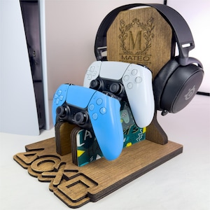 May include: Wooden gaming console stand with a headset, two controllers, and the word "PlayStation" cut out. The stand has a personalized wooden back with the name "Mateo" and the letter "M". One controller is blue, the other is white.