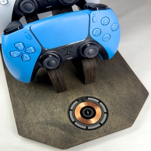 May include: A blue and black game controller rests on a dark wooden stand with a wireless charging pad. The stand has a unique shape and supports the controller. Another controller is visible in the background.
