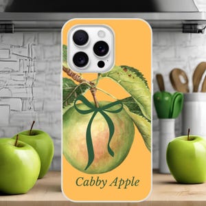 Puede incluir: Funda de teléfono con una ilustración de acuarela de una manzana verde con un lazo verde oscuro y hojas. La funda tiene un fondo amarillo y el texto "Cabby Apple".