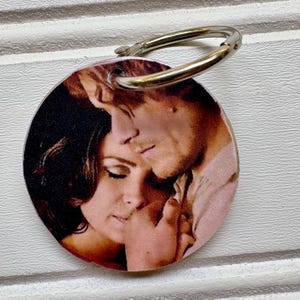 May include: Round keychain featuring a romantic image of a man and woman embracing. The image is printed on a circular tag with a silver-colored metal ring for attachment.