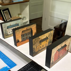 Four primitive Americana decor blocks displayed together on a white shelf in a craft room, featuring vintage colonial saltbox homes in distressed blue, red, cream, and mustard tones with early American farmhouse style.