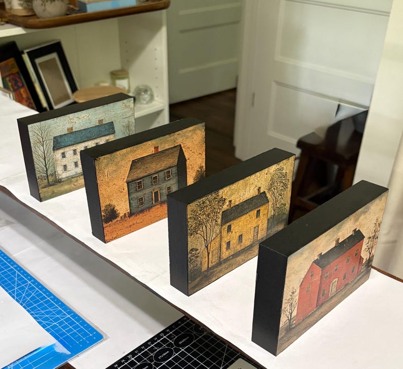 Four primitive Americana decor blocks displayed together on a white shelf in a craft room, featuring vintage colonial saltbox homes in distressed blue, red, cream, and mustard tones with early American farmhouse style.