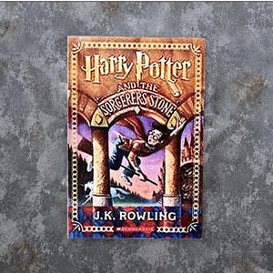 May include: A rectangular book cover for Harry Potter and the Sorcerer's Stone by J.K. Rowling. The cover features a central illustration of a figure flying on a broomstick between two stone pillars, with a unicorn visible in the lower left corner.