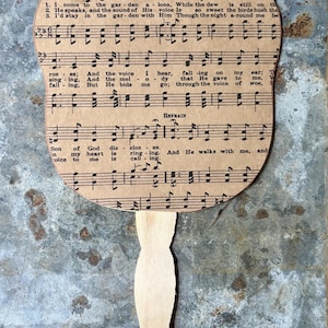 May include: A vintage-style paper fan with musical notes and lyrics from "In the Garden." The fan features a light brown handle and a curved top with printed sheet music. The text includes the title and lyrics.