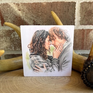 May include: A square wooden block featuring a detailed color illustration of a woman with curly brown hair wearing a plaid shawl and a man with wavy hair in a dark jacket, with the man's hand gently touching the woman's face in a close embrace.
