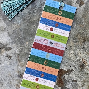 May include: A colorful bookmark featuring the book titles by Diana Gabaldon, with a teal tassel. The titles include "Outlander," "Voyager," and "Dragonfly in Amber." The bookmark is a vertical rectangle.