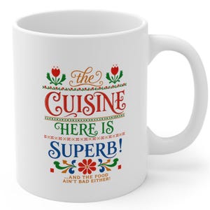 May include: A white ceramic mug featuring a colorful graphic design with the text "the CUISINE HERE IS SUPERB! ...AND THE FOOD AIN'T BAD EITHER!" surrounded by red, green, and blue floral and geometric patterns, including two red tulips at the top.
