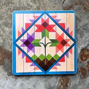 May include: A square wooden coaster featuring a printed geometric quilt-style pattern. The design displays a central flower motif in red, purple, and green, enclosed within a blue diamond border on a light wood grain background with vertical lines.